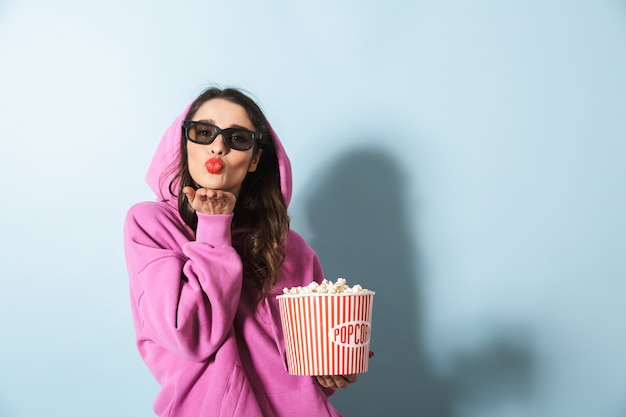 Retrato de mujer joven positiva con gafas 3D sonriendo mientras sostiene el cubo de palomitas de maíz sobre azul en estudio