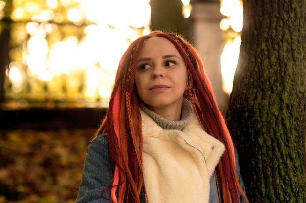 Retrato de mujer joven positiva con rastas de jengibre mirando alrededor de pie cerca del tronco del árbol