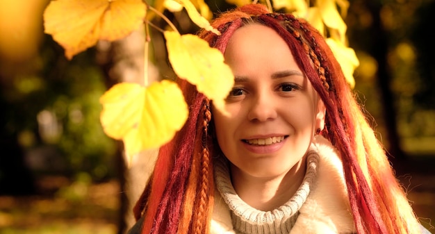 Retrato de mujer joven positiva con rastas de jengibre de pie entre ramas de árbol con hojas amarillas