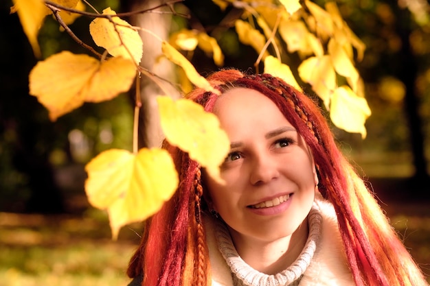 Retrato de mujer joven positiva con rastas de jengibre de pie entre ramas de árbol con hojas amarillas