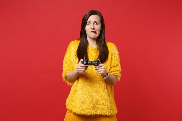 Retrato de mujer joven preocupada en suéter de piel amarilla mordiendo los labios, jugando videojuegos con joystick aislado sobre fondo rojo brillante. Personas sinceras emociones, concepto de estilo de vida. Simulacros de espacio de copia.