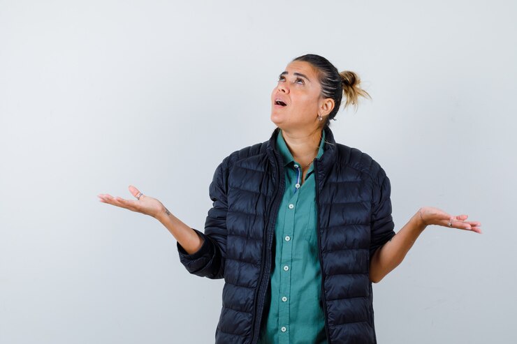 Foto retrato de mujer joven que muestra un gesto de impotencia, mirando hacia arriba en camisa, chaqueta acolchada y mirando pensativo vista frontal