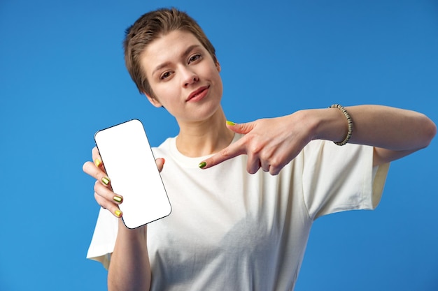 Retrato de una mujer joven que muestra un teléfono móvil con pantalla en blanco mientras está de pie sobre un fondo azul