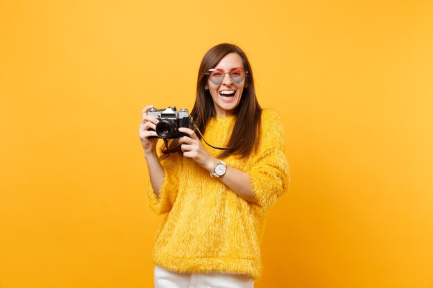 Retrato de mujer joven riendo en anteojos de corazón tomando fotografías en la cámara de fotos vintage retro aislada sobre fondo amarillo brillante. Personas sinceras emociones, concepto de estilo de vida. Área de publicidad.