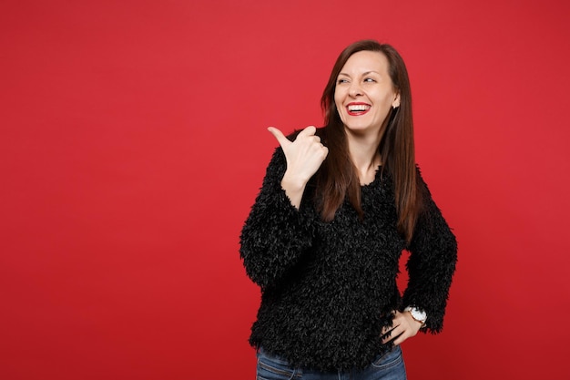 Retrato de mujer joven riendo en pie de suéter de piel negra, apuntando con el pulgar a un lado aislado sobre fondo rojo brillante de la pared en el estudio. Personas sinceras emociones, concepto de estilo de vida. Simulacros de espacio de copia.