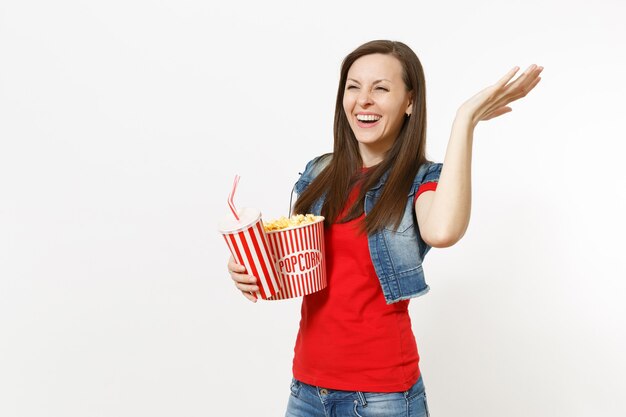 Retrato de mujer joven riendo en ropa casual viendo una película, sosteniendo un cubo de palomitas de maíz, un vaso de plástico de refresco o cola extendiendo la mano aislada sobre fondo blanco. Emociones en el concepto de cine.