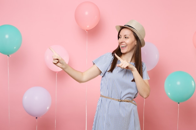 Retrato de mujer joven riendo con sombrero de paja de verano y vestido azul apuntando con el dedo índice a un lado sobre fondo rosa con coloridos globos de aire. Fiesta de cumpleaños, concepto de emociones sinceras de personas.