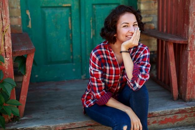 Retrato de mujer joven rizada cerca de casa vieja al aire libre en un día soleado. Niña sonriente y emocional en jeans y camisa roja en la calle de la ciudad.