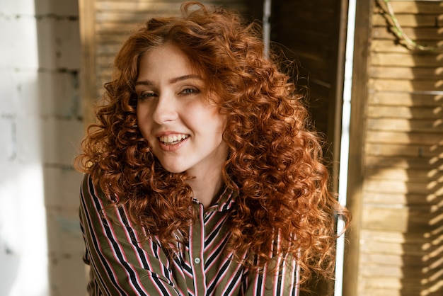 Retrato de mujer joven rizada pelirroja feliz cerca de la ventana sonriendo closeup