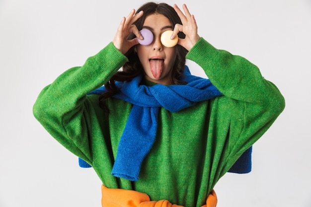 Retrato de mujer joven en ropa colorida sosteniendo galletas de macarrón, mientras está de pie aislado en blanco