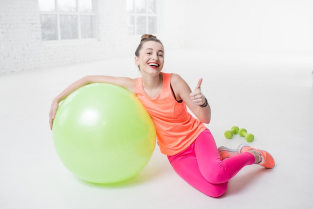 Retrato de una mujer joven en ropa deportiva colorida con pelota de fitness en el suelo en el gimnasio blanco