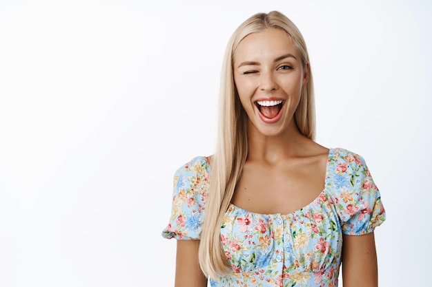 Retrato de mujer joven rubia guiñando un ojo sonriendo y luciendo entusiasta de pie en vestido sobre fondo blanco.