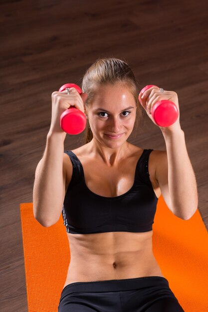 Foto retrato de mujer joven rubia sonriente haciendo abdominales con pesas de mano sobre alfombra naranja brillante y mirando a cámara en estudio de ejercicio