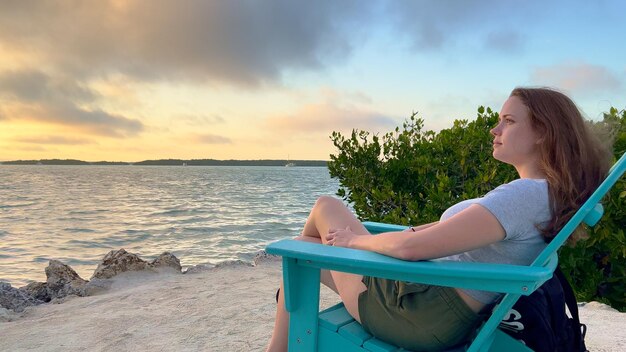 Retrato de una mujer joven sentada en la playa contra el cielo durante la puesta de sol