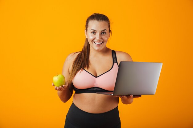 Retrato de una mujer joven con sobrepeso feliz vistiendo ropa deportiva que se encuentran aisladas sobre la pared amarilla, usando la computadora portátil, sosteniendo la manzana verde