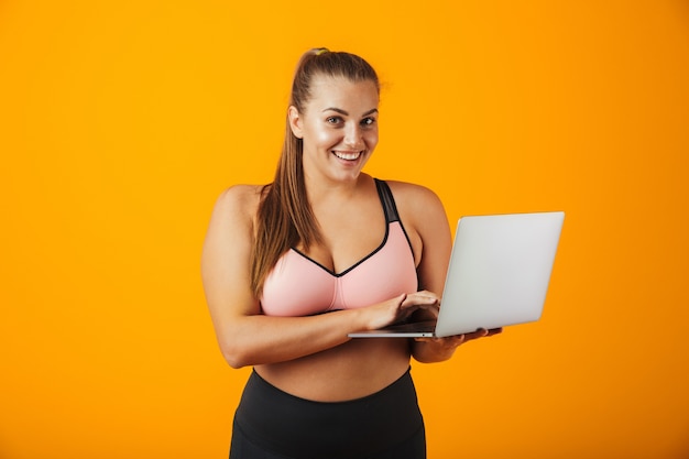 Retrato de una mujer joven con sobrepeso feliz vistiendo ropa deportiva que se encuentran aisladas sobre la pared amarilla, utilizando equipo portátil