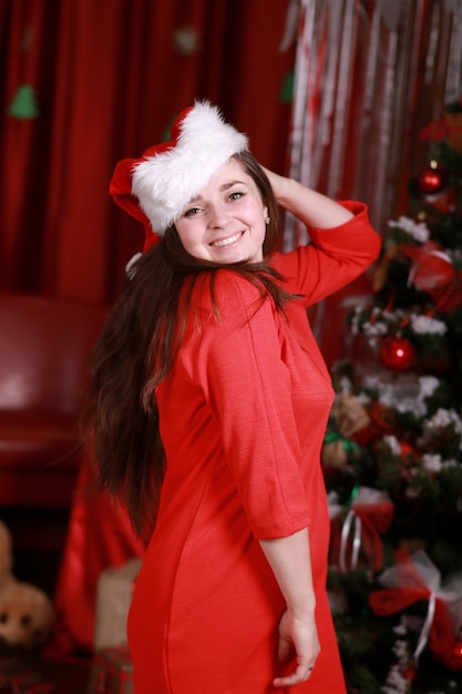 Retrato de una mujer joven con sombrero de ayudante de santa y vestido rojo en preparación para la Navidad en casa. Navidad, Navidad, invierno, concepto de felicidad - mujer sonriente en la pared del árbol de brillo de Navidad.