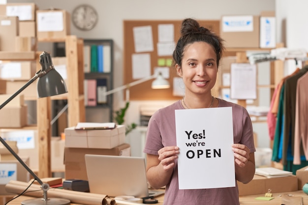 Retrato de mujer joven sonriendo al frente sosteniendo cartel mientras está de pie en la oficina