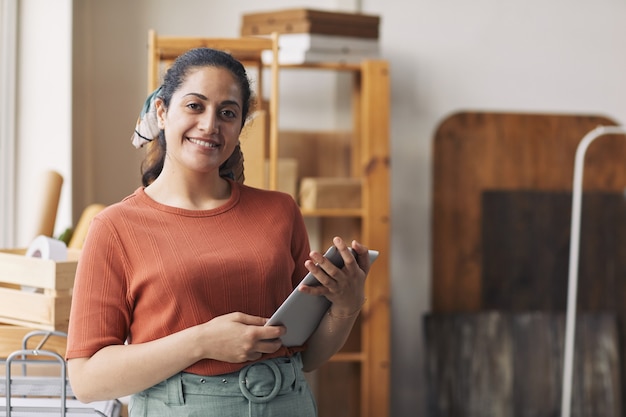 Retrato de mujer joven sonriendo a la cámara mientras usa tableta digital durante su trabajo en el almacén