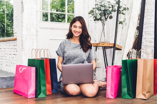 Retrato de una mujer joven sonriendo usando una computadora portátil mientras está sentada junto a las bolsas de compras en casa