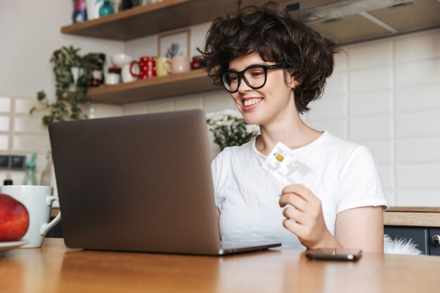 Retrato de una mujer joven sonriente con anteojos trabajando en la computadora portátil en casa por la mañana