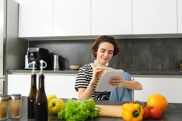 Foto retrato de una mujer joven sonriente en la cocina sosteniendo un cuaderno haciendo notas para escribir recetas de comestibles