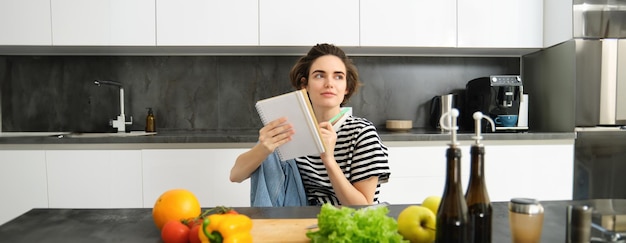 Retrato de una mujer joven sonriente en la cocina sosteniendo un cuaderno haciendo notas para escribir recetas de comestibles