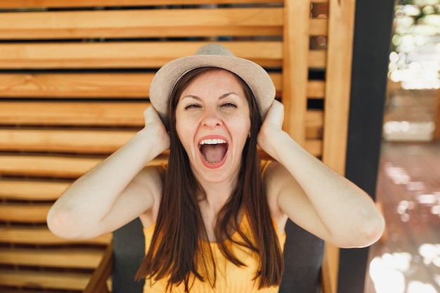 Retrato de mujer joven sonriente emocionada con sombrero de paja de verano, camisa amarilla puso las manos en la cabeza sobre la pared de madera en la calle al aire libre cafetería de verano café