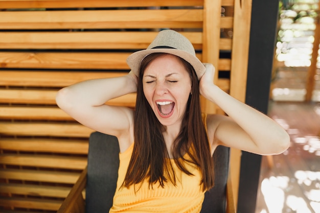 Retrato de mujer joven sonriente emocionada con sombrero de paja de verano, camisa amarilla puso las manos en la cabeza sobre la pared de madera en la calle al aire libre cafetería de verano café