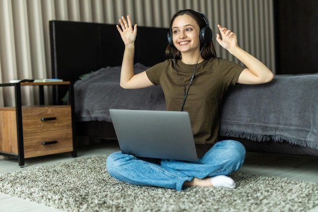Retrato de una mujer joven sonriente escuchando música con auriculares y usando la computadora portátil mientras se apoya en un sofá en casa