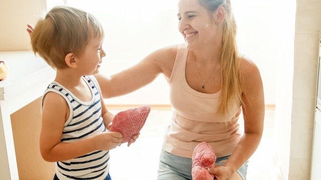 Retrato de mujer joven sonriente feliz con 3 años de edad, niño pequeño hijo de pie en la cocina contra la ventana grande