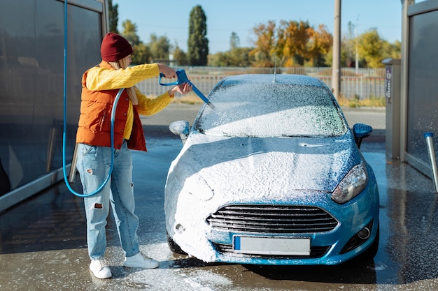 Retrato mujer joven, sonriente, feliz y atractiva lavando automóviles en la estación de autoservicio de lavado manual de automóviles, limpieza con espuma, agua a presión. Transporte, automóvil, concepto de cuidado de vehículos.