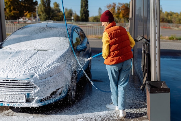 Retrato mujer joven, sonriente, feliz y atractiva lavando automóviles en la estación de autoservicio de lavado manual de automóviles, limpieza con espuma, agua a presión. Transporte, automóvil, concepto de cuidado de vehículos.