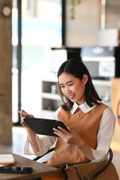 Retrato de mujer joven sonriente navegar navegando por internet inalámbrico en tableta digital mientras está sentado en la cafetería.