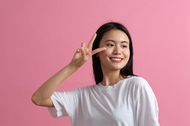 Retrato de mujer joven sonriente positiva asiática con gesto de dedos V sobre fondo rosa.