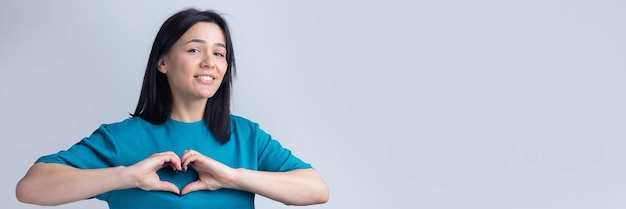 Retrato de una mujer joven sonriente que muestra un gesto de corazón con los dedos aislado sobre gris