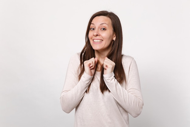 Retrato de una mujer joven y sonriente con ropa ligera mirando la cámara y apretando los puños aislados en el fondo de la pared blanca en el estudio. Emociones sinceras de la gente, concepto de estilo de vida. Simulacros de espacio de copia.