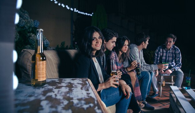 Retrato de mujer joven sonriente sentada y sosteniendo una cerveza mientras sus amigos hablando en una fiesta al aire libre. Concepto de amistad y celebraciones.