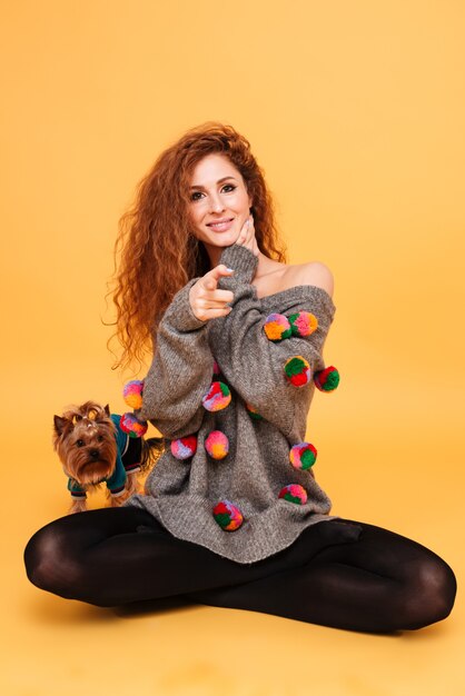 Foto retrato de una mujer joven sonriente sentada con su perrito yorkshire terrier y señalar con el dedo aislado en la pared naranja