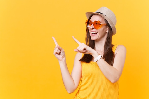 Retrato de mujer joven sonriente con sombrero de paja de verano, gafas naranjas apuntando con el dedo índice a un lado copia espacio aislado sobre fondo amarillo. Personas sinceras emociones, concepto de estilo de vida. Área de publicidad.