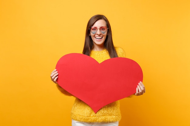 Retrato de mujer joven sonriente en suéter de piel y anteojos de corazón con corazón rojo en blanco vacío aislado sobre fondo amarillo brillante. Personas sinceras emociones, concepto de estilo de vida. Área de publicidad.