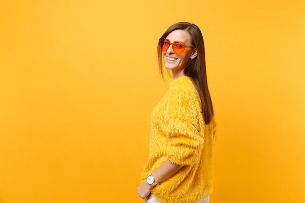 Retrato de mujer joven sonriente en suéter de piel, pantalón blanco y gafas de corazón naranja mirando hacia atrás aislado sobre fondo amarillo brillante. Personas sinceras emociones, concepto de estilo de vida. Área de publicidad.