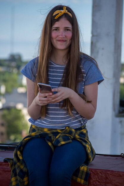 Retrato de una mujer joven sonriente usando un teléfono móvil mientras está sentada en una pared de contención