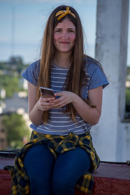 Foto retrato de una mujer joven sonriente usando un teléfono móvil mientras está sentada en una pared de contención