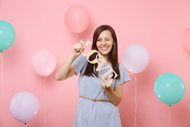 Retrato de mujer joven sonriente en vestido azul parpadeando sosteniendo letras de madera palabra amor sobre fondo rosa pastel con coloridos globos de aire. Fiesta de cumpleaños, concepto de emociones sinceras de personas.