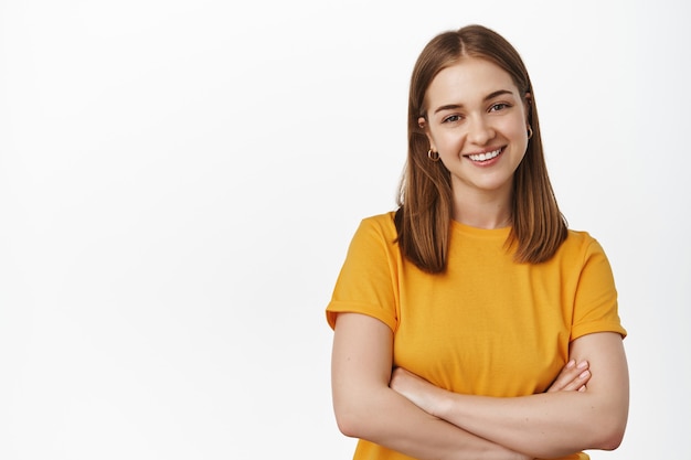 Retrato de mujer joven con sonrisa segura, pose profesional relajada, vistiendo camiseta amarilla de empresa, brazos cruzados en el pecho, de pie contra la pared blanca