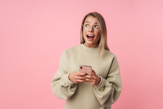 Retrato de mujer joven sorprendida en anteojos con teléfono celular y mirando hacia arriba aislado sobre pared rosa