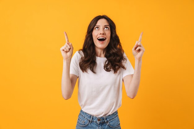 Retrato de mujer joven sorprendida feliz emocional posando aislada sobre pared amarilla apuntando.