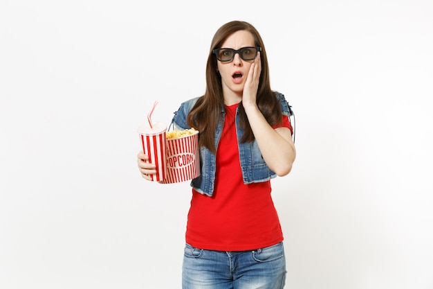 Retrato de mujer joven sorprendida en gafas 3d viendo una película, sosteniendo un cubo de palomitas de maíz y un vaso de plástico de refresco o cola, manteniendo la mano cerca de la cara aislada sobre fondo blanco. Emociones en el cine.