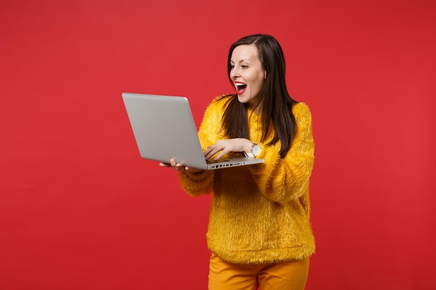 Retrato de mujer joven sorprendida en suéter de piel amarilla manteniendo la boca abierta usando una computadora portátil aislada sobre fondo rojo brillante. Personas sinceras emociones, concepto de estilo de vida. Simulacros de espacio de copia.